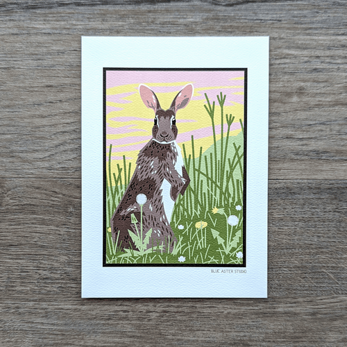 A 5x7 art print of a rabbit standing alert in a grassy area surrounded by dandelions and clover. There is a pink and yellow sunrise behind the rabbit which gives a little glow to its ears.