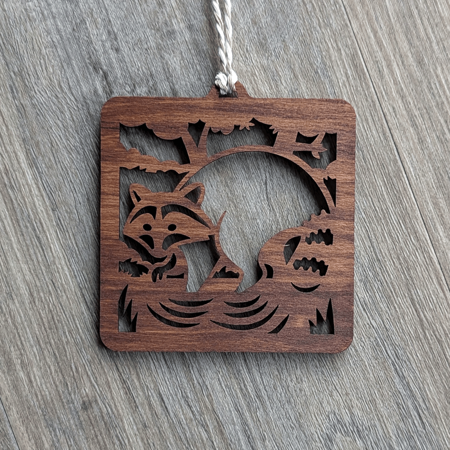 A square brown wooden ornament of a raccoon in a stream.