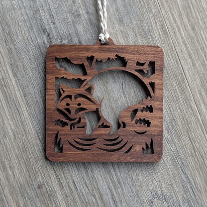 A square brown wooden ornament of a raccoon in a stream.