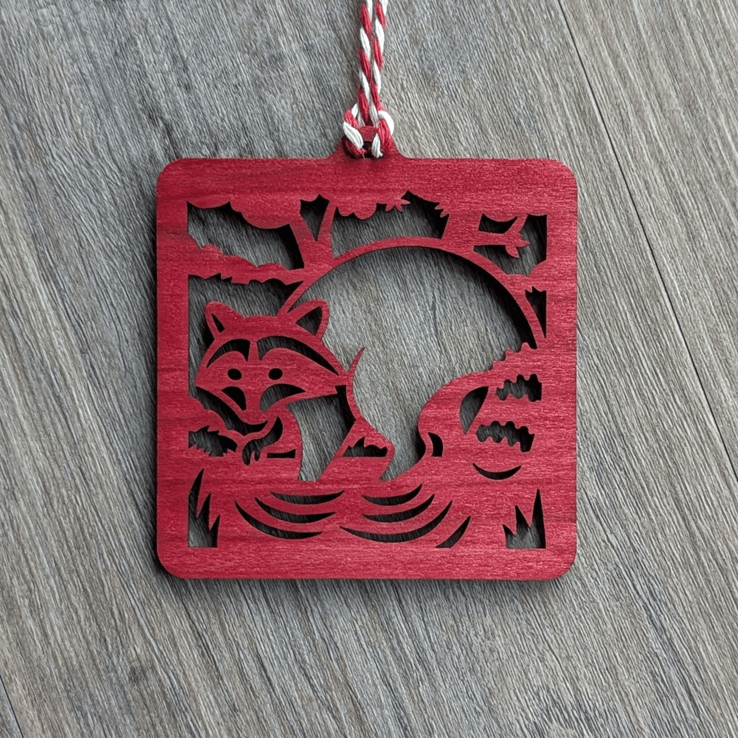 A square red wooden ornament of a raccoon in a stream.