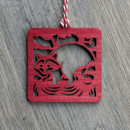 A square red wooden ornament of a raccoon in a stream.