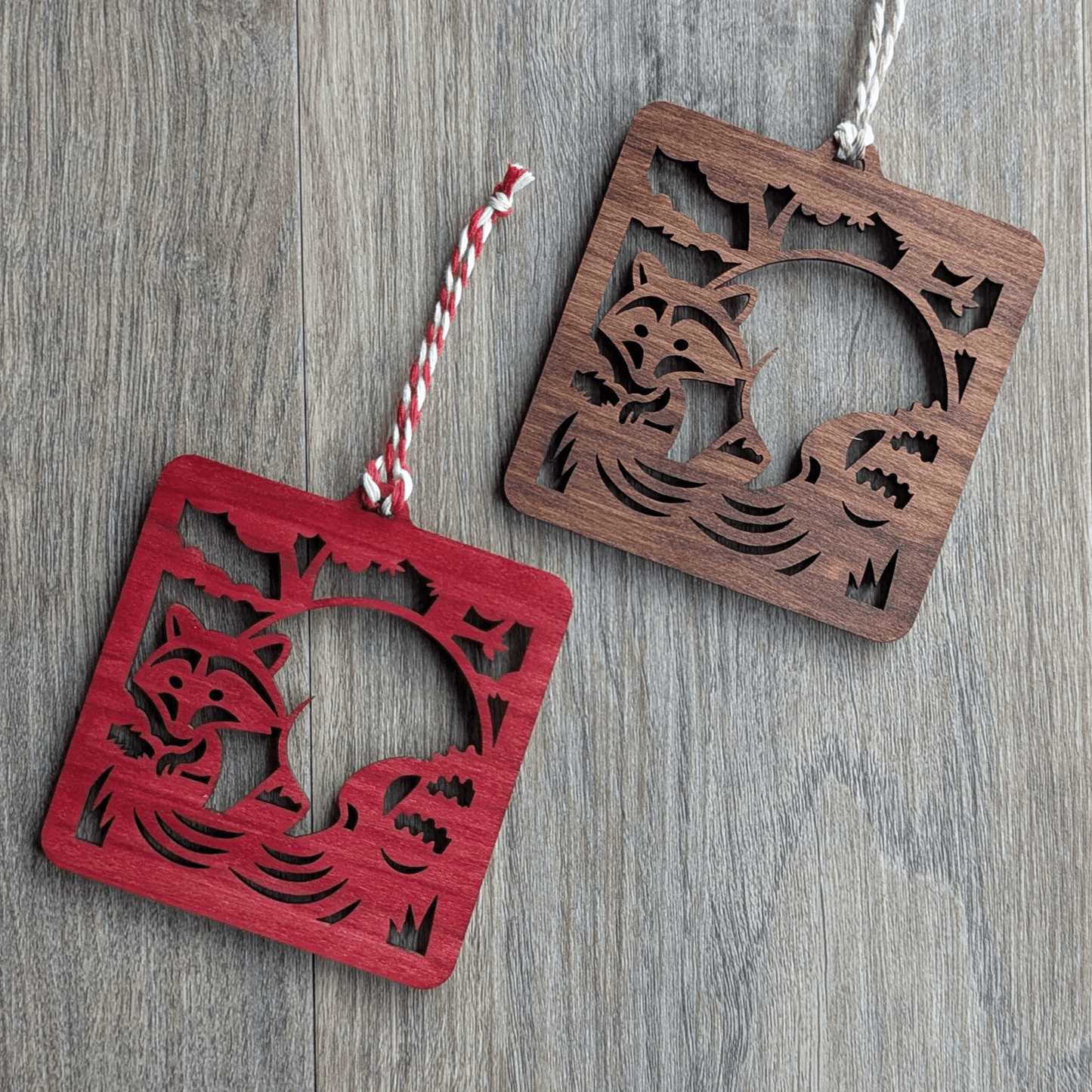 Two square wooden ornaments of a raccoon in a stream.