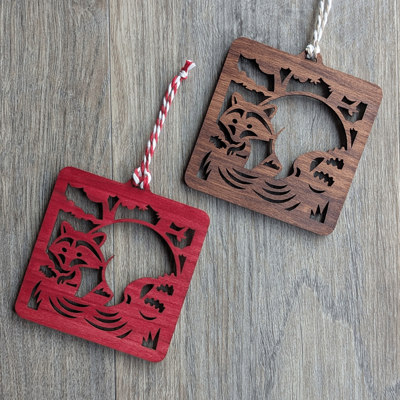 Two square wooden ornaments of a raccoon in a stream.