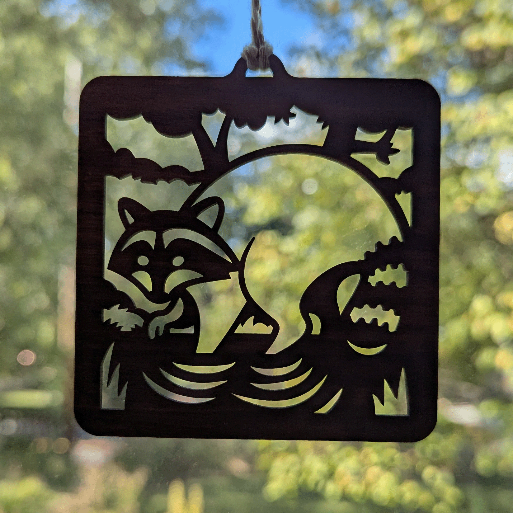 A square wooden ornament of a raccoon in a stream hanging in a window.