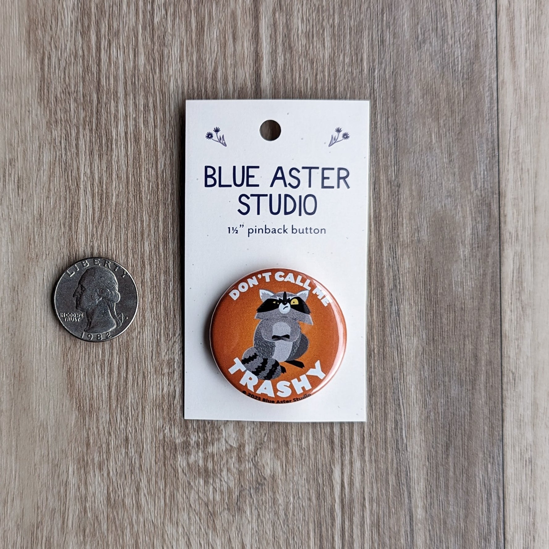 A one and a half inch pinback button featuring a raccoon with its arms crossed and an annoyed expression on its face with an orange background. The text reads "Don't call me trashy." A quarter is beside the button to show scale.