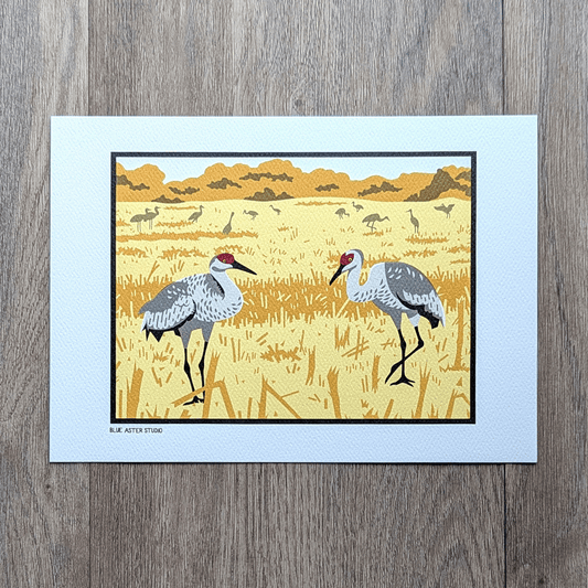 An illustrated print of a pair of sandhill cranes, large gray birds with red patches on their heads, in a stubbly field of corn. A flock of birds in silhouette are in the distance.