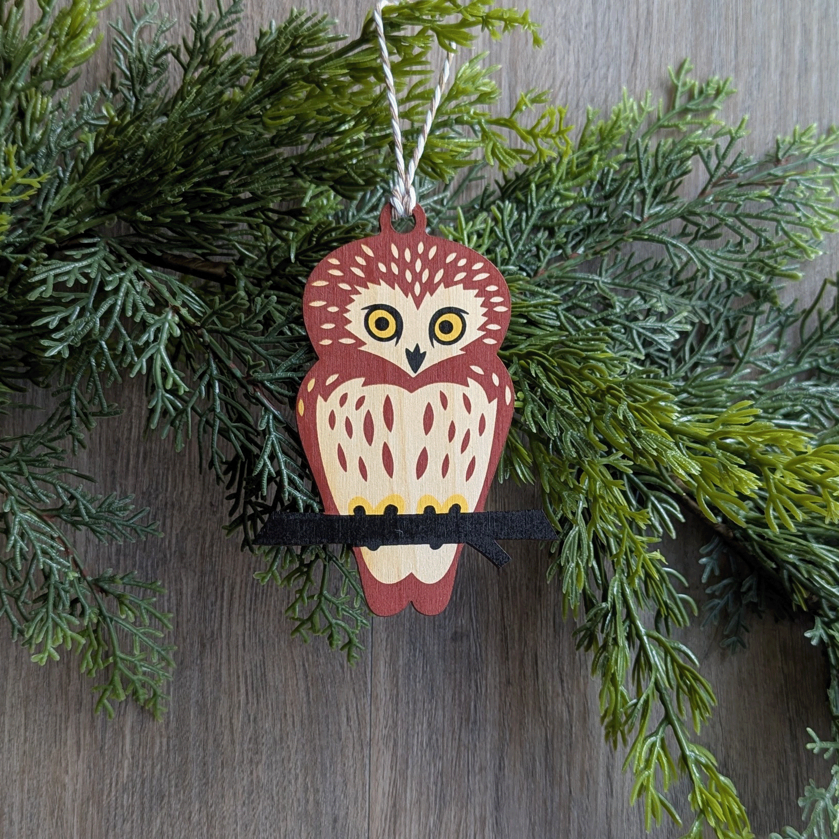 A wooden screen printed ornament of a saw-whet owl hanging on a garland.
