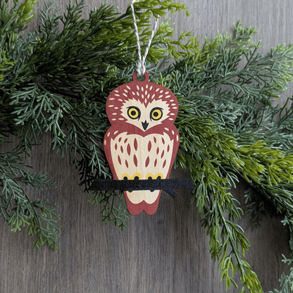 A wooden screen printed ornament of a saw-whet owl hanging on a garland.