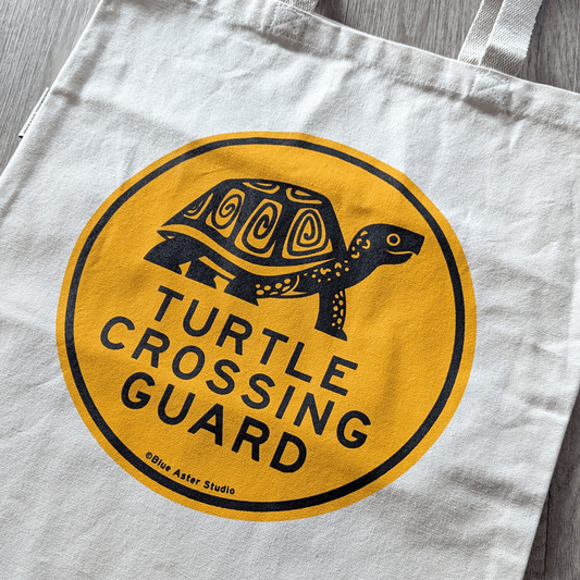 A sturdy canvas tote bag with a yellow circle with the words Turtle Crossing Guard and an illustration of a box turtle.