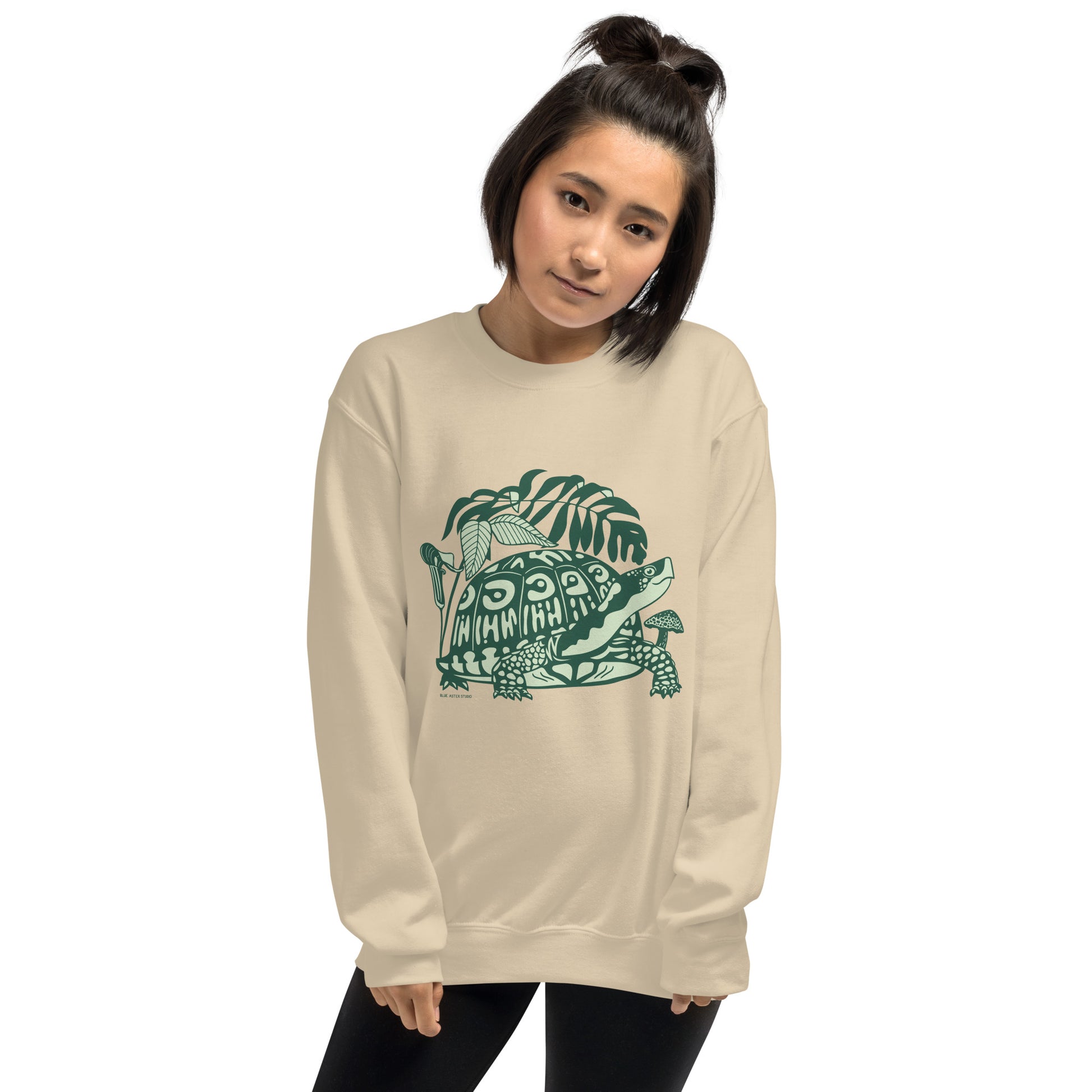 A woman wearing a light beige crew neck sweatshirt with a design of a box turtle surrounded by woodland plants.