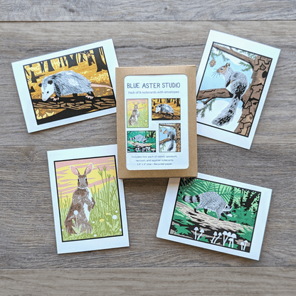 A collection of eight wildlife notecards featuring an illustration of an opossum walking on a log in the woods, a squirrel sitting on an evergreen branch, a rabbit standing in a grassy field, and a raccoon walking on a log on a forest floor. The pack includes two cards of each design plus envelopes.