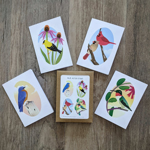 A package of notecards with a four notecards arranged around it. The cards each feature an illustration of a birds: an eastern bluebird, an American goldfinch, a male and female American cardinal, and a ruby-throated hummingbird.