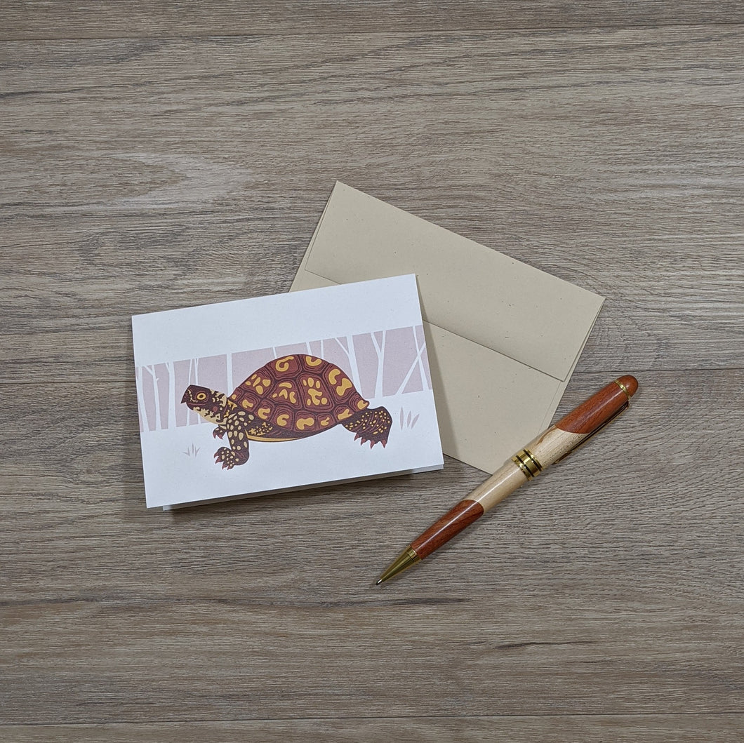 A note card with an illustration of a box turtle. The card is siting on the natural speckled brown envelope and next to it is a pen.