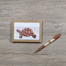 Load image into Gallery viewer, A package of 8 box turtle cards and envelopes shown in their kraft paper box with an open window which reveals the design. Next to the note card pack is a pen.