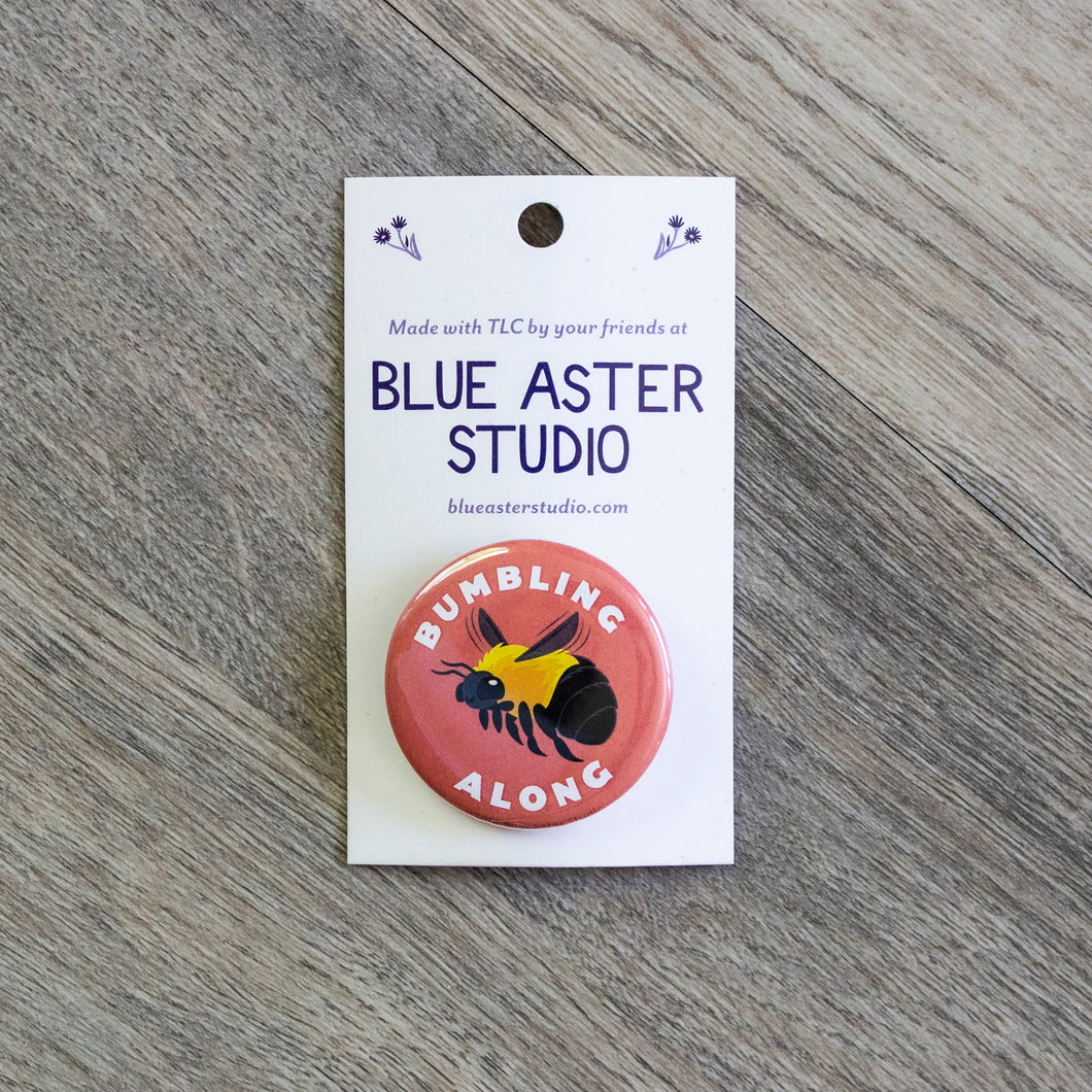 A 1.5 inch pink pinback button with an illustration of a bumble bee and the words Bumbling Along around it