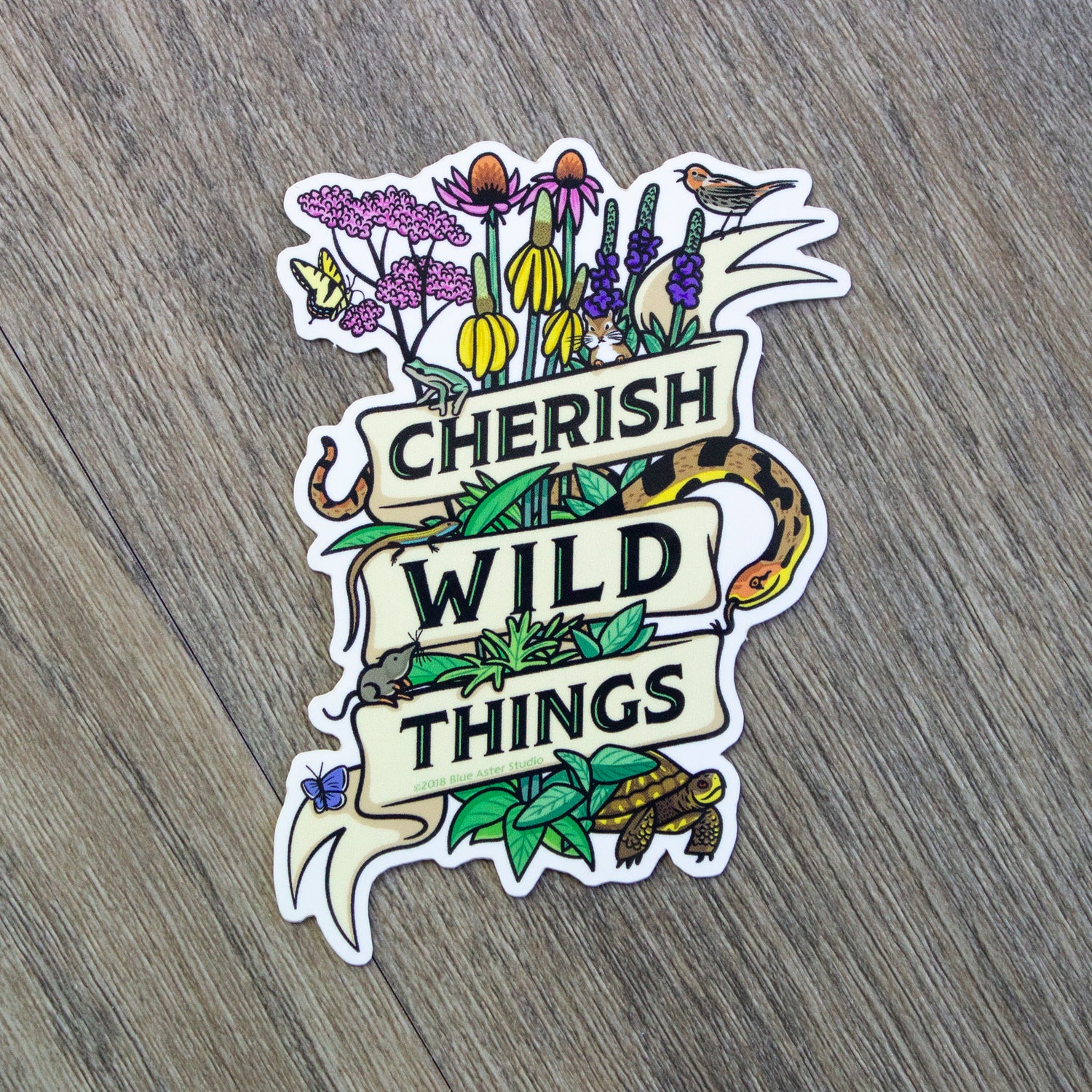 A vinyl sticker of a collection of prairie plants and animals wrapped up with a ribbon that reads Cherish Wild Things.