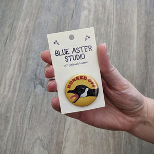 A hand holding the honked off goose button that is on a backing card with the Blue Aster Studio logo on it.