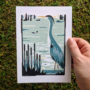 A hand holding an art print of a heron in a swampy wetland.