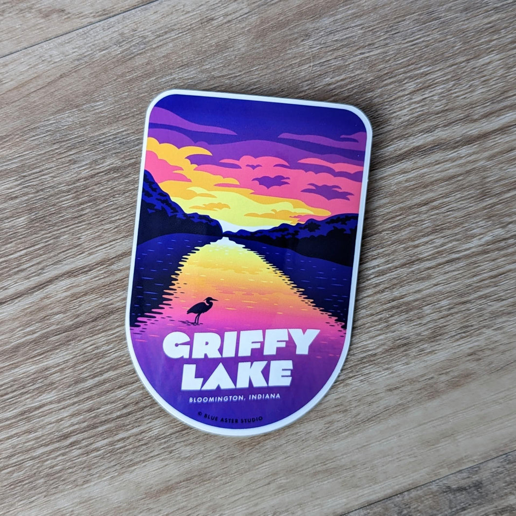 A shield shaped vinyl sticker that features an illustration of a sunset over Griffy Lake with a silhouette of a heron in the foreground. At the bottom of the sticker there is text that says Griffy Lake Bloomington, Indiana.