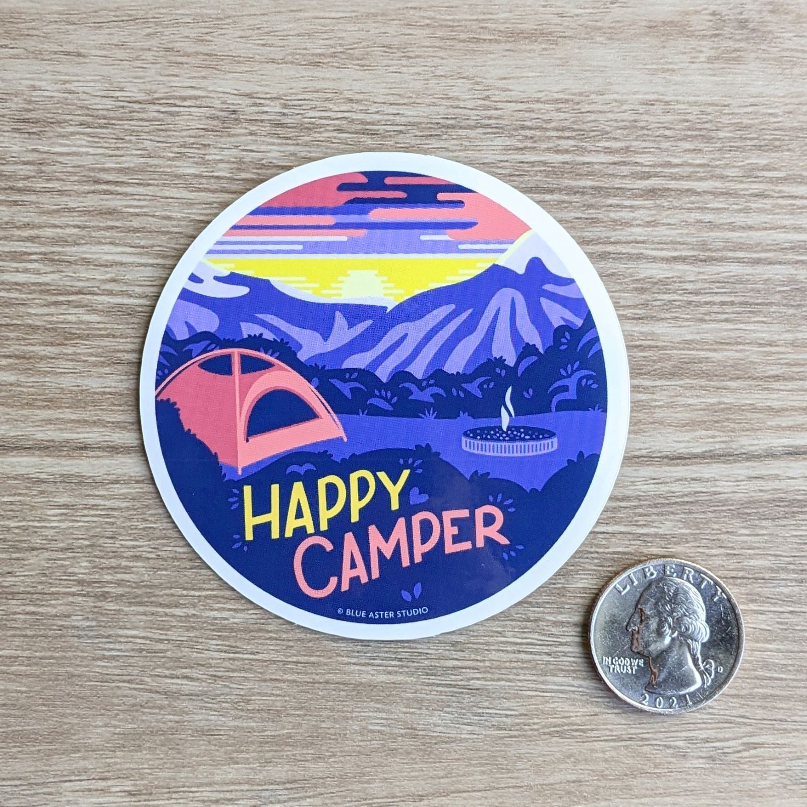 A round vinyl sticker depicting a campsite in the mountains at sunrise with the text "Happy Camper" with a quarter to show scale. The sticker measures three inches in diameter.