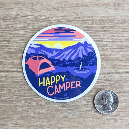 A round vinyl sticker depicting a campsite in the mountains at sunrise with the text "Happy Camper" with a quarter to show scale. The sticker measures three inches in diameter.