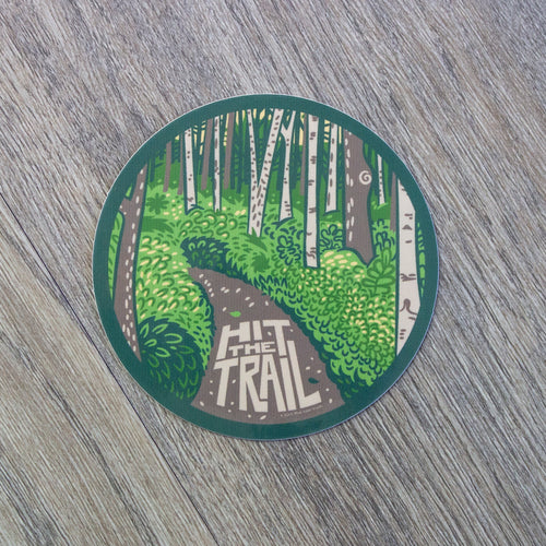 A round vinyl sticker with a forest path scene with the words Hit The Trail leading down the path.