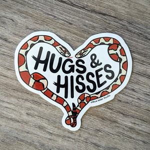 A vinyl sticker featuring illustrations of two milk snakes that are forming a heart around the words "Hugs & Hisses"