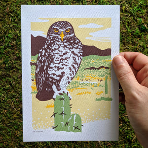 A hand holding an 5x7 inch art print of a burrowing owl in a desert scene.