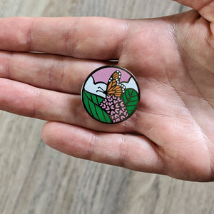 A hand holding a circular enamel pin featuring a monarch butterfly nectaring on a common milkweed flower.