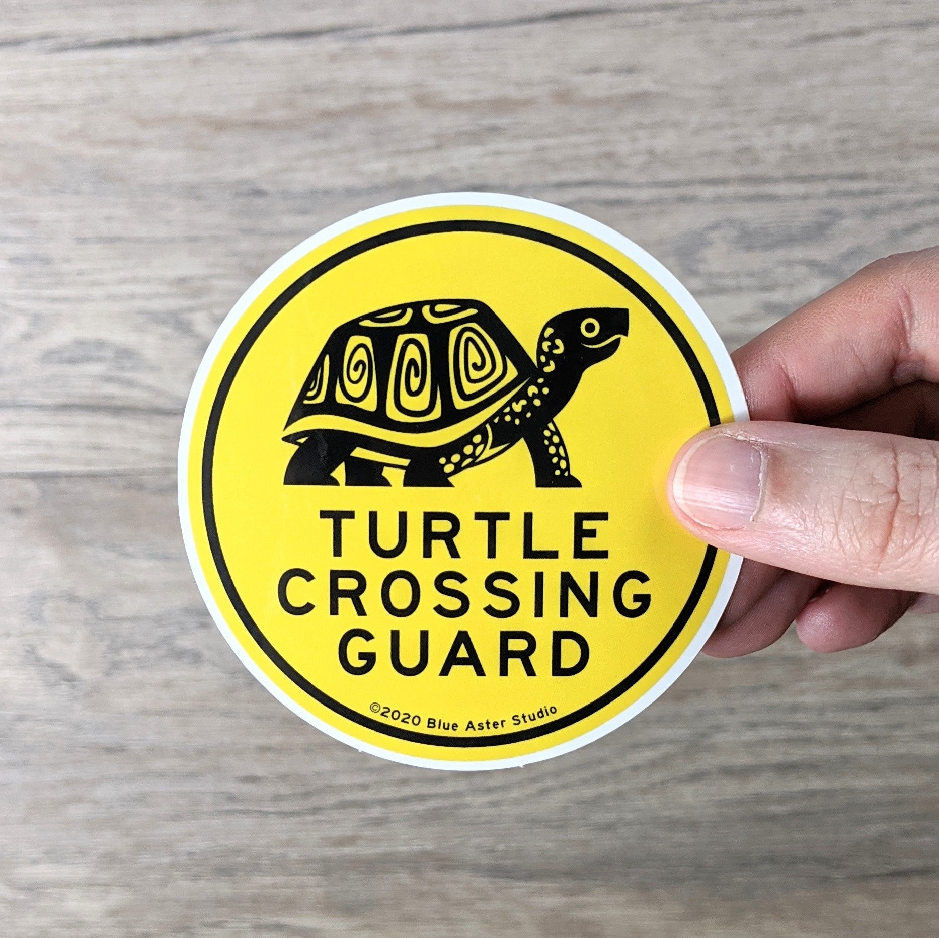 Hand holding a 3 inch round, yellow vinyl sticker with an illustration of a box turtle and the words "Turtle Crossing Guard" in black.