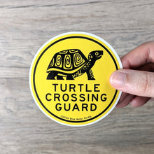 Load image into Gallery viewer, Hand holding a 3 inch round, yellow vinyl sticker with an illustration of a box turtle and the words &quot;Turtle Crossing Guard&quot; in black.