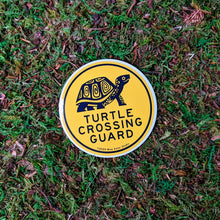 Load image into Gallery viewer, A round 3 inch vinyl sticker with an illustration of a box turtle and the words &quot;Turtle Crossing Guard&quot; in black on yellow. The sticker is sitting on a mossy background.