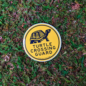 A round 3 inch vinyl sticker with an illustration of a box turtle and the words "Turtle Crossing Guard" in black on yellow. The sticker is sitting on a mossy background.