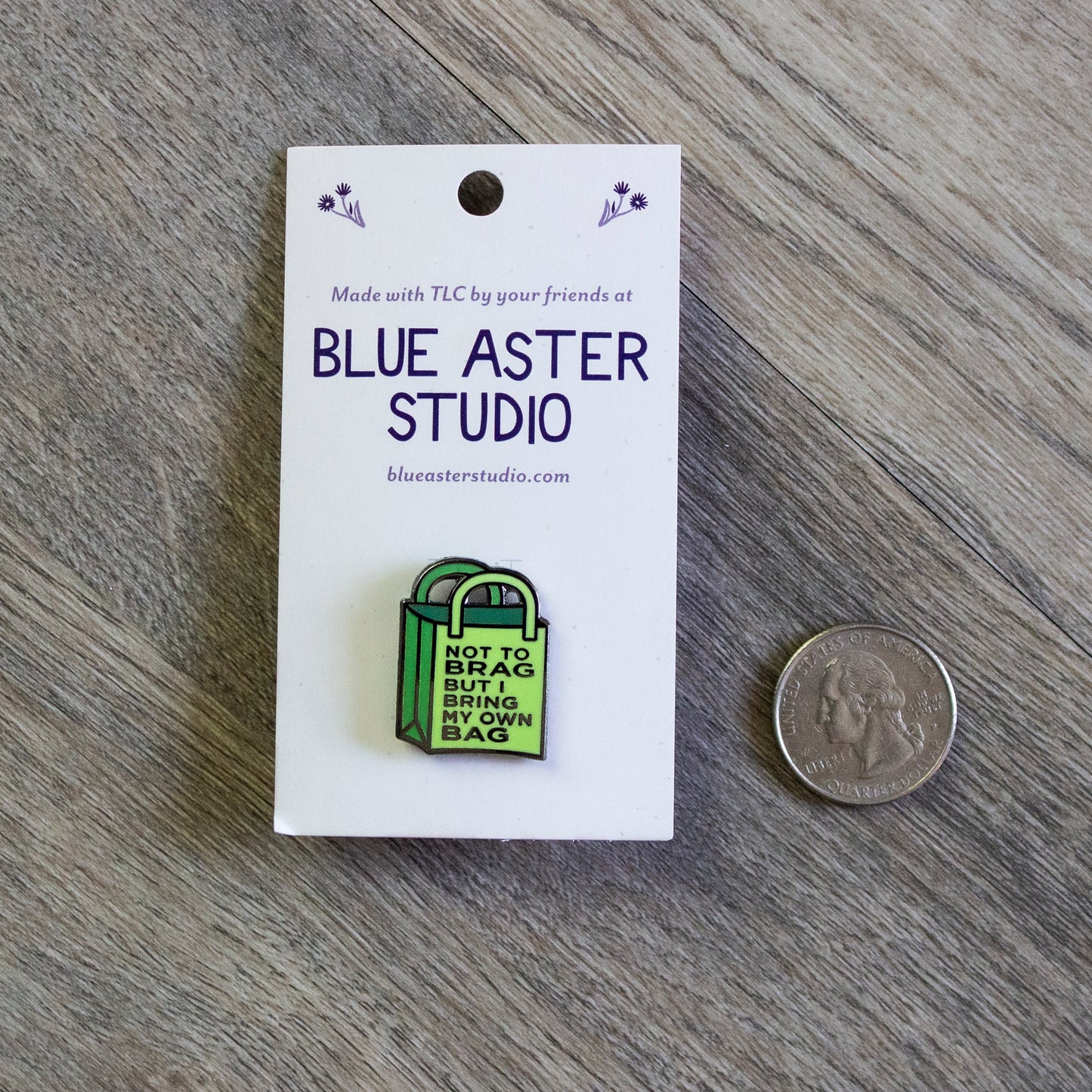 The Not To Brag But I Bring My Own Bag enamel pin sitting next to a USD quarter for scale.