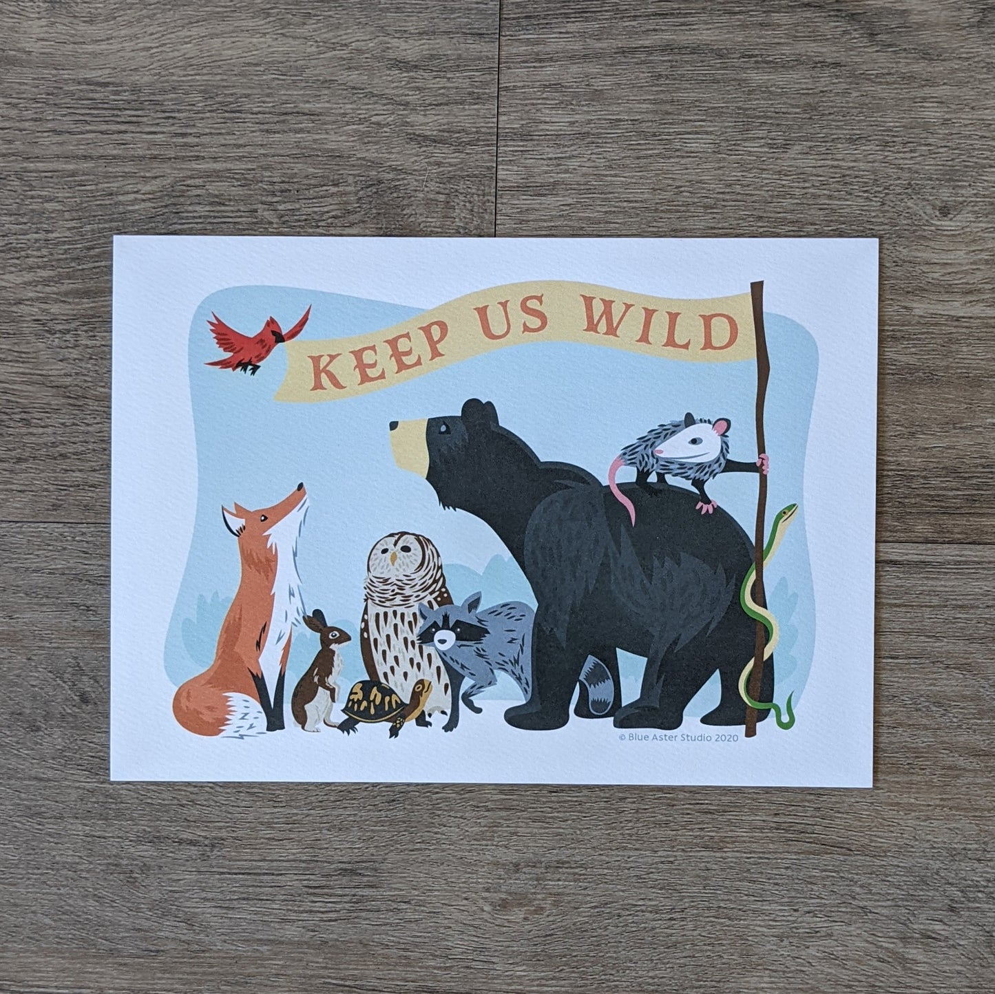 An art print of an illustration of a group of animals (bear, opossum, snake, raccoon, owl, turtle, rabbit, fox, and bird) holding a flag that reads "Keep Us Wild"