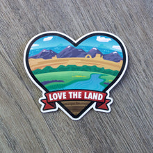 Load image into Gallery viewer, A heart shaped vinyl sticker with a ribbon across the bottom with the words Love The Land. Within the heart is a landscape scene that spans from prairie to the mountains to the sea.