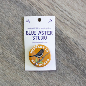 A 1.5 inch orange pinback button with an illustration of a Mockingbird perched on a branch and the words Prepare To Be Mocked.