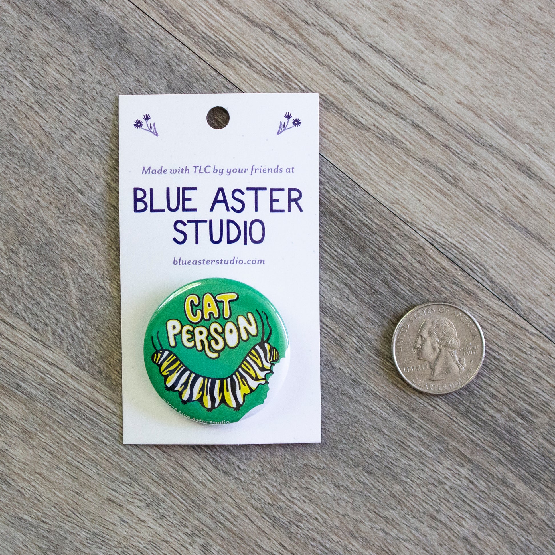A 1.5 inch green pinback button with an illustration of a monarch caterpillar crawling along the bottom and the words Cat Person above it. A USD quarter is provided for scale.