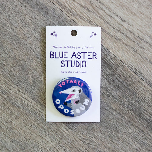 A 1.5 inch blue pinback button with an illustration of an opossum and the words Totally Opossum.