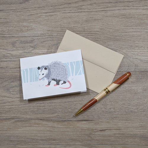 A card featuring an opossum illustration sits on top of a natural speckled brown envelope with a pen to the side.