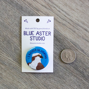 A pinback button with an illustration of an osprey and the words "I'd Rather Be Fishing" sitting next to a USD quarter for scale.