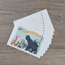 Load image into Gallery viewer, A stack of ten &quot;Keep Us Wild&quot; postcards with an illustration of a fox, rabbit, turtle, owl, raccoon, bear, opossum, snake, and songbird all holding a banner with the words &quot;Keep Us Wild&quot; on it.