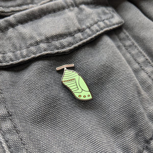 A monarch chrysalis enamel pin on a gray canvas jacket. The pin is photographed at an angle to show the shine of the metal.
