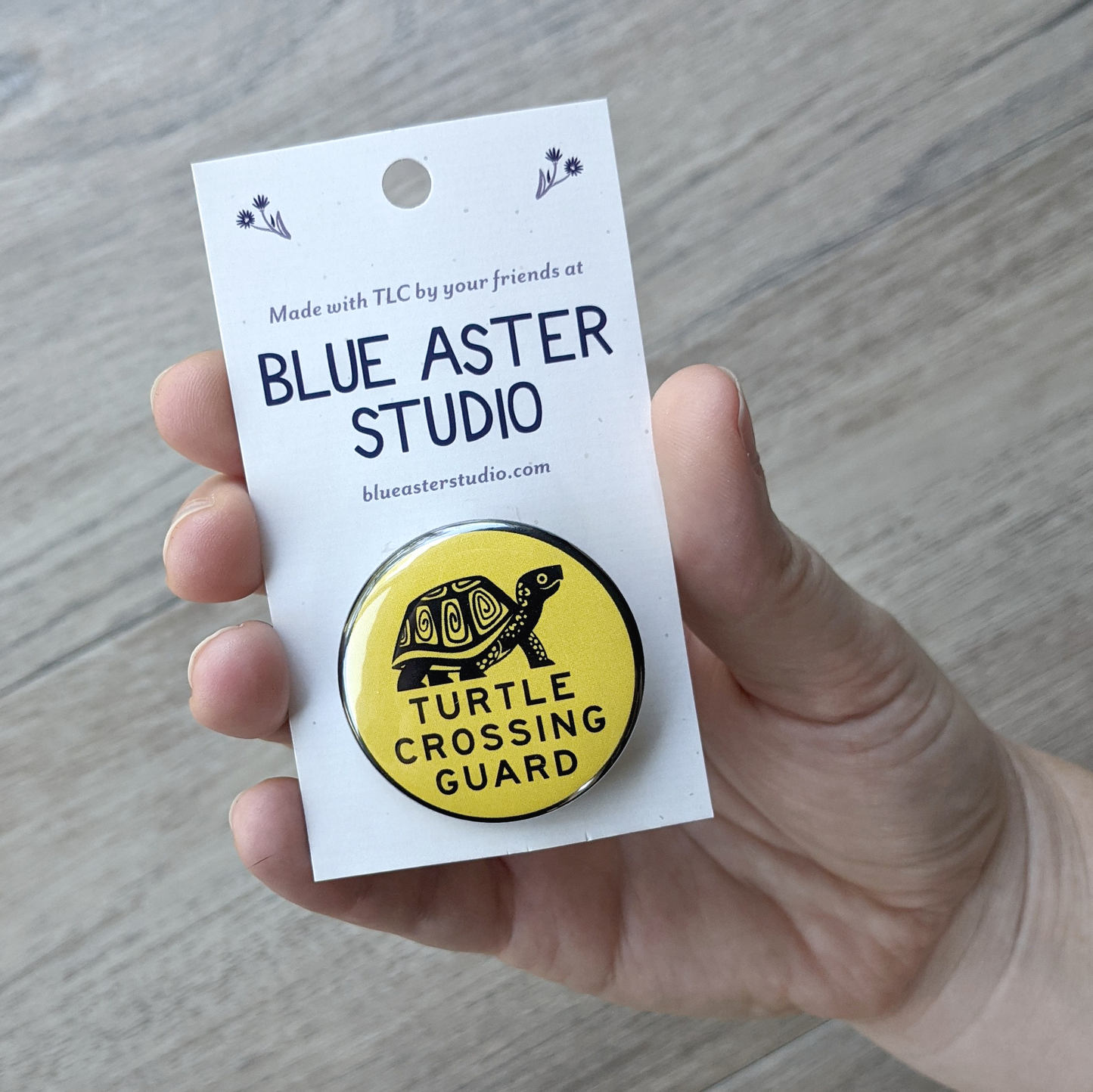 A hand holding a 1.5 inch pinback button on a backer card. The button has an illustration of a box turtle and the words "Turtle Crossing Guard" in black on a yellow background.