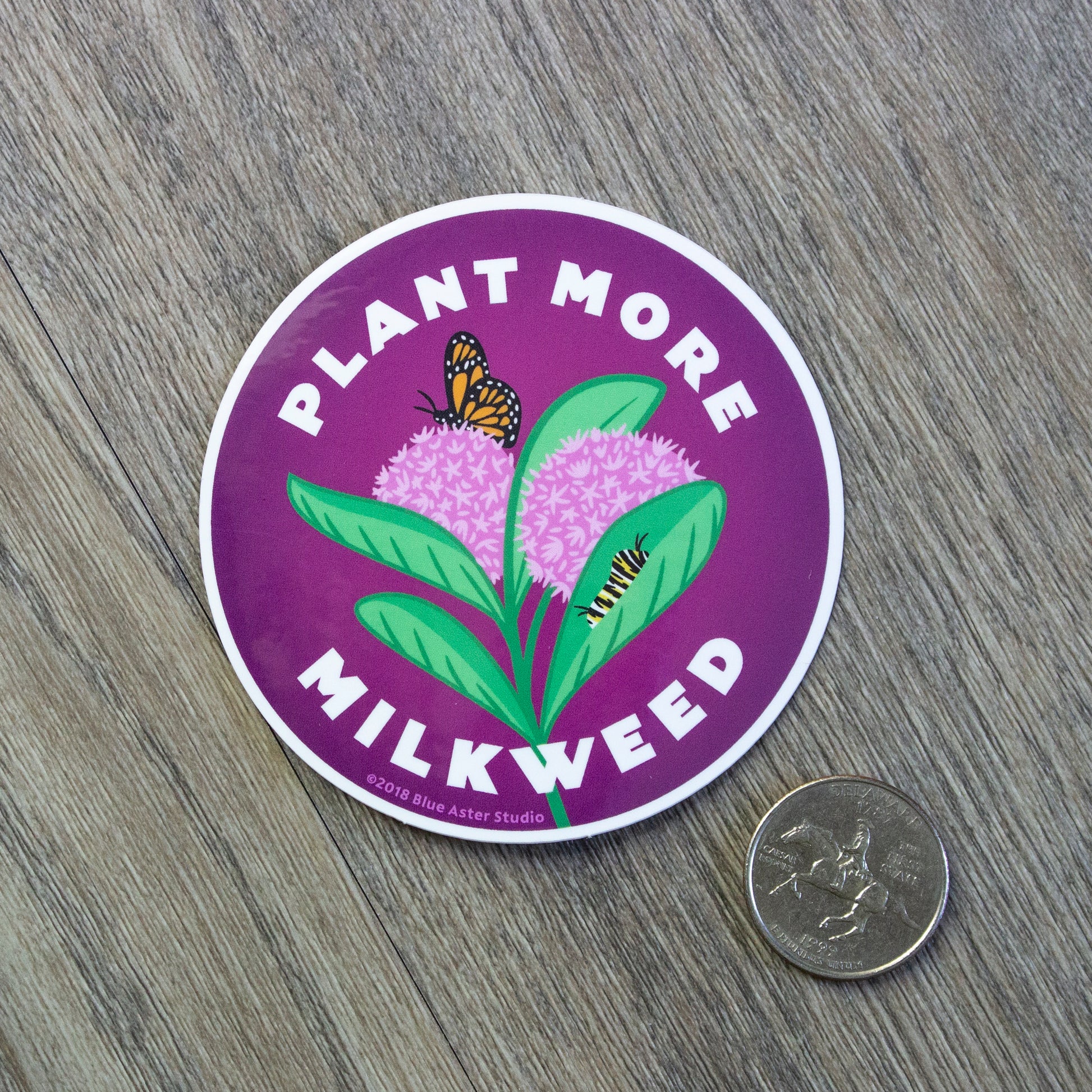 A round vinyl sticker with an illustration of a common milkweed plant in the center and the words Plant More Milkweed around it. The milkweed sticker is sitting next to a USD quarter for scale.
