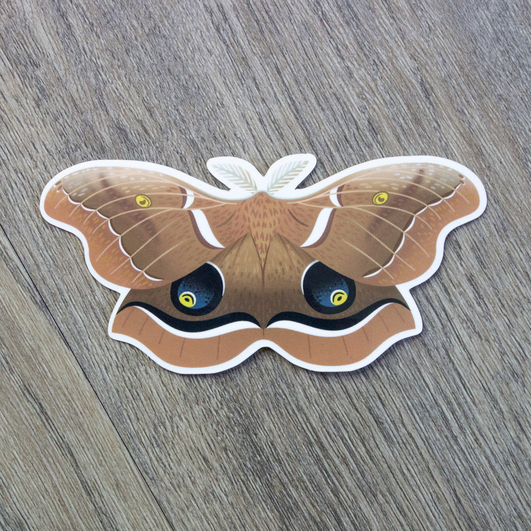 A vinyl sticker of a polyphemus moth. The sticker is sitting on a wood background.