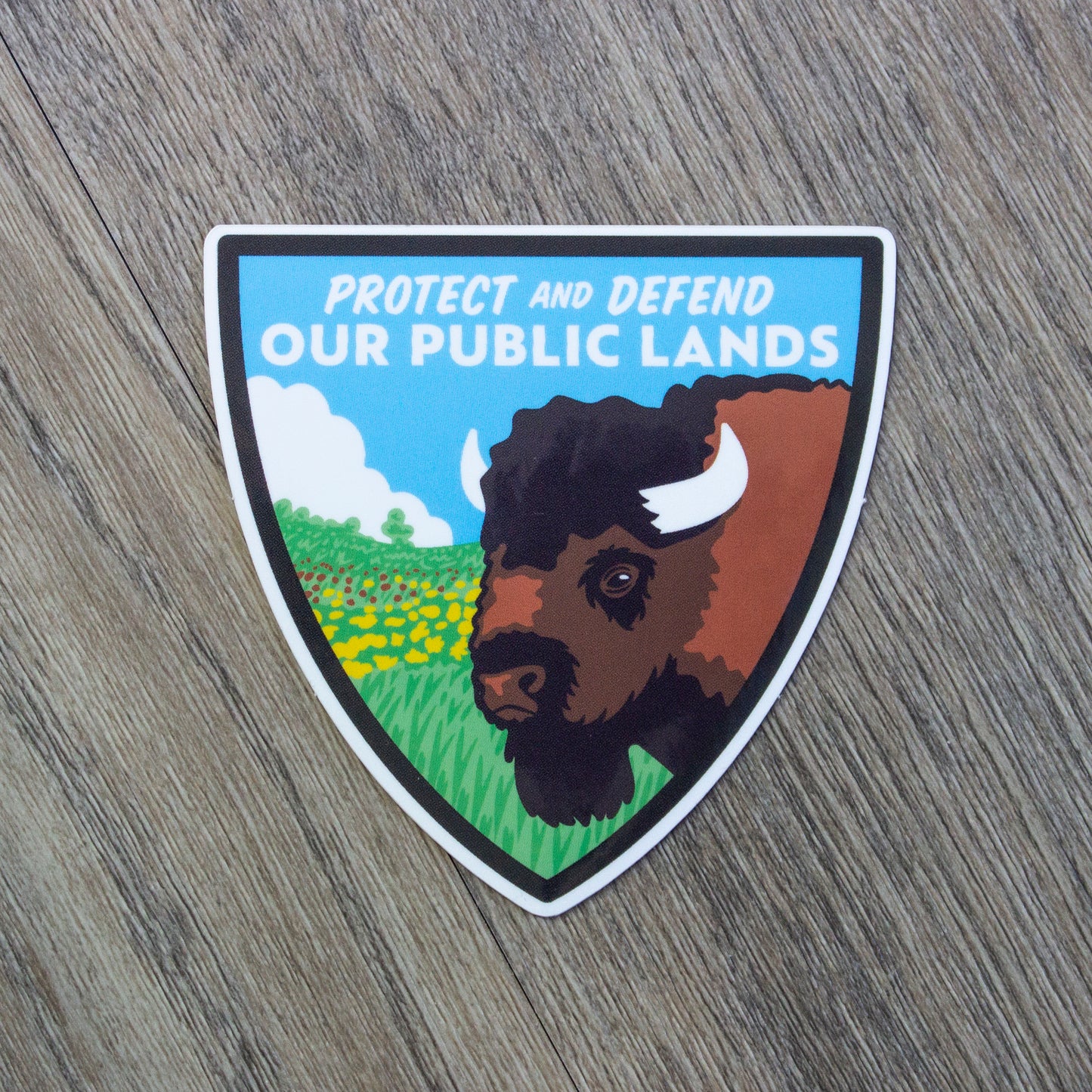 A shield shaped vinyl sticker with a bison and prairie scene and the words Protect And Defend Our Public Lands at the top.