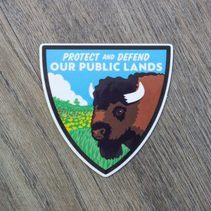A shield shaped vinyl sticker with a bison and prairie scene and the words Protect And Defend Our Public Lands at the top.