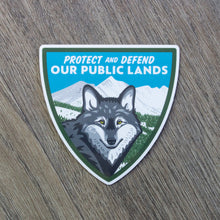Load image into Gallery viewer, A shield shaped vinyl sticker with a wolf and mountain scene and the words Protect And Defend Our Public Lands at the top.