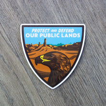 Load image into Gallery viewer, A shield shaped vinyl sticker with an eagle and desert scene and the words Protect And Defend Our Public Lands at the top.